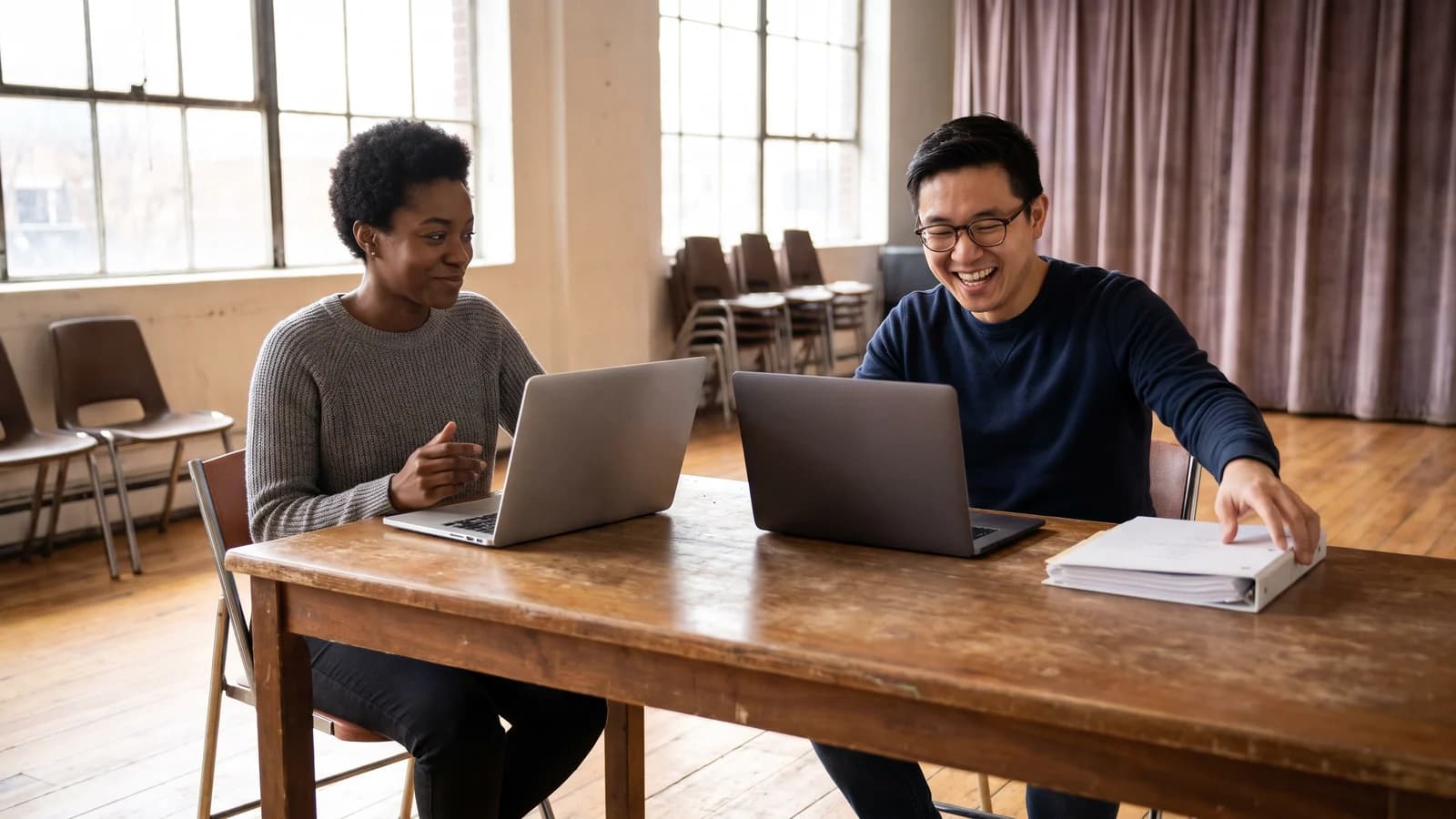Two screenwriters using different unbranded laptops while shaping the same scene together in a bright rehearsal room